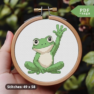 May include: A cross-stitch pattern of a green frog waving with its right hand. The pattern is in a wooden embroidery hoop and the text "Stitches: 49 x 58" is below the hoop.
