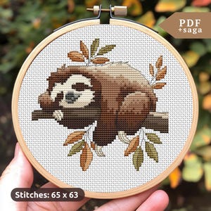 Puede incluir: Bordado de punto de cruz de un perezoso descansando en una rama, rodeado de hojas. El perezoso es marrón y beige, con una cara clara. El bordado está en un aro de madera. Texto: "PDF + saga" y "Stitches: 65 x 63".
