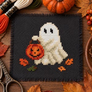 May include: A square black fabric piece featuring a cross-stitch design of a white ghost holding an orange jack-o'-lantern bucket. The ghost is surrounded by small orange and green leaf motifs. The fabric has a frayed, fringed edge finish.