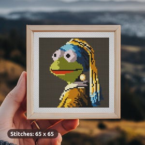 May include: A framed cross-stitch of a green frog wearing a blue and yellow patterned headscarf and a pearl earring. The frog is looking to the right. The cross-stitch is 65 stitches wide by 65 stitches tall.
