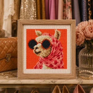 May include: A square cross-stitch embroidery in a light wood frame featuring a llama wearing dark round sunglasses and a pink patterned scarf against a vibrant orange background. The needlework displays a detailed animal portrait with visible stitches.