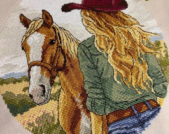 May include: A cross-stitch embroidery featuring a person with long blonde hair wearing a dark red cowboy hat, a green shirt, and blue jeans, standing beside a brown and white horse with a bridle. The text CherryMarryStore is visible.