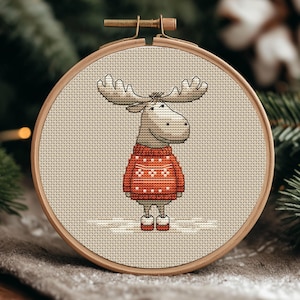 Puede incluir: Un bastidor de bordado de punto de cruz con un alce de dibujos animados que lleva un suéter rojo con patrones de copos de nieve blancos. El alce tiene grandes astas y está sobre un fondo neutro.