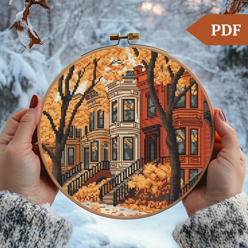 May include: Cross stitch pattern of a row of houses with autumn leaves on the ground. The houses are white and red with brown trim and windows. The pattern is in a wooden embroidery hoop.