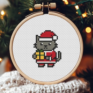 May include: A cross-stitch embroidery hoop featuring a gray cat wearing a Santa hat and suit, holding a wrapped gift. The cat is centered on a white background with a wooden hoop. The background is blurred with Christmas tree branches and lights.