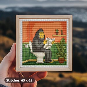 Puede incluir: Obra de arte de punto de cruz en un marco de madera. La imagen representa un gorila sentado en un inodoro, leyendo un periódico. El fondo presenta una pared naranja, baldosas verdes y plantas en macetas. El texto "Stitches: 65 x 65" está en la parte inferior.