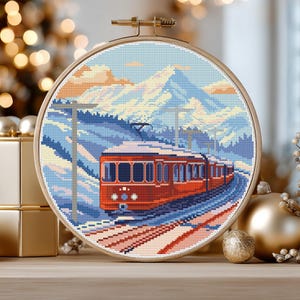 Puede incluir: Un bordado de punto de cruz en un aro de madera representa un tren rojo que viaja por un paisaje montañoso nevado. La obra de arte presenta un cielo azul, montañas cubiertas de nieve y un tren detallado. El aro se exhibe sobre una superficie de madera.