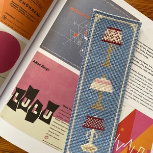 Actual photo of the cross-stitch bookmark, stitched in bright blue and resting on an open art book.