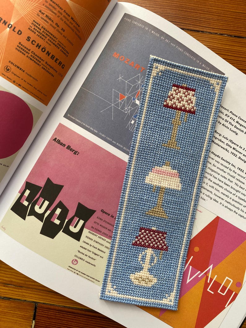 Actual photo of the cross-stitch bookmark, stitched in bright blue and resting on an open art book.