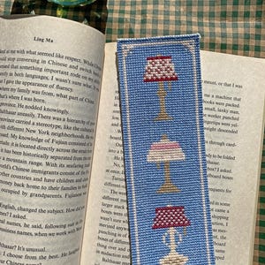 Actual photo of the cross-stitch bookmark, stitched in bright blue and resting on the open page of a book in the sunlight