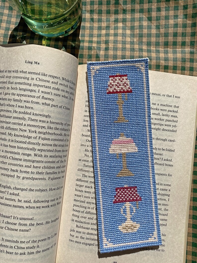 Actual photo of the cross-stitch bookmark, stitched in bright blue and resting on the open page of a book in the sunlight