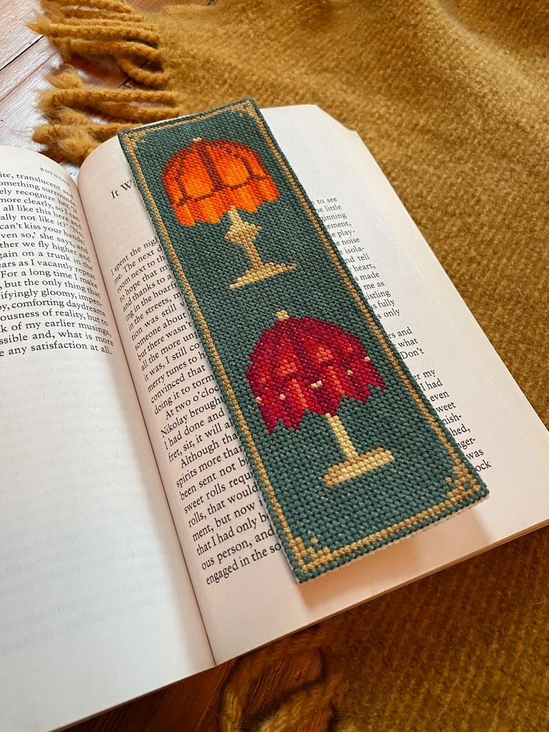 Close up angle of the stitched example of Fancy Lamps cross stitch bookmark pattern finished, resting inside a book on top of antique hard wood floors.