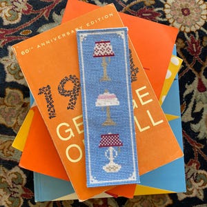 Actual photo of the cross-stitch bookmark, stitched in bright blue and resting on a stack of orange and blue books.