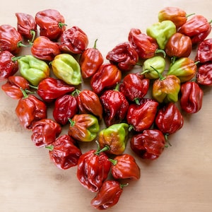 May include: A heart-shaped arrangement of red and green habanero peppers on a light brown wooden surface. The peppers vary in color from bright red to green and yellow, with a textured, wrinkled appearance.