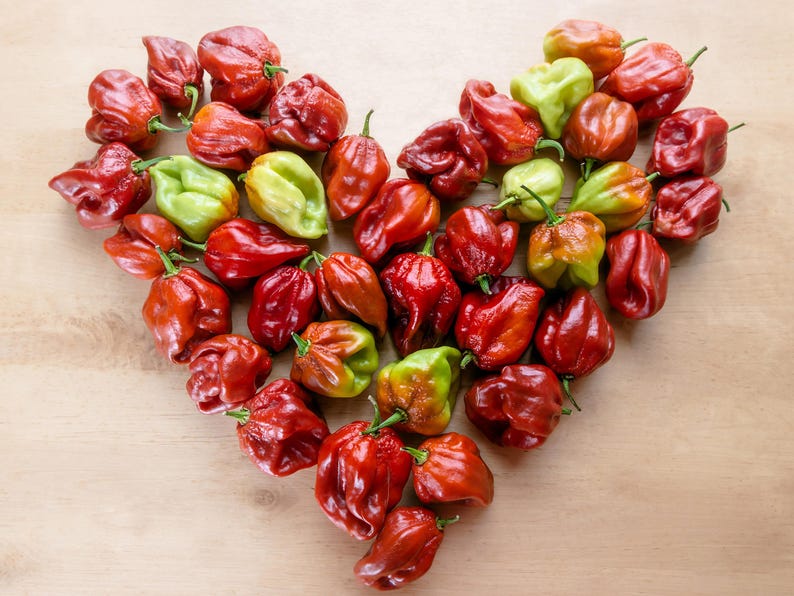 May include: A heart-shaped arrangement of red and green habanero peppers on a light brown wooden surface. The peppers vary in color from bright red to green and yellow, with a textured, wrinkled appearance.