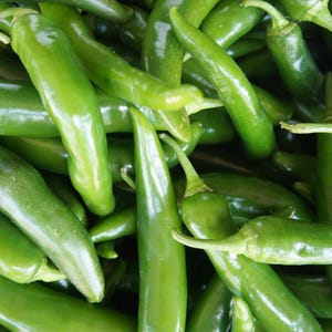 May include: Close-up of a pile of fresh, vibrant green chili peppers. The peppers have a smooth, glossy texture and a long, slender shape. The image showcases the natural color and texture of the peppers, perfect for culinary use.