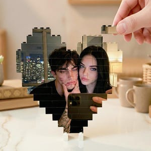May include: A heart-shaped photo display made of black blocks, featuring a selfie of a couple. The background shows a city skyline. A hand is placing a block on the top of the heart. The display sits on a white surface.