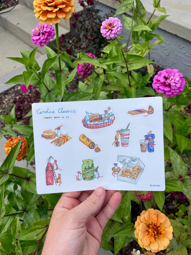 May include: A white sticker sheet featuring illustrations of North Carolina food items, including a hot dog, burger, fries, soda, and donuts. The text "Carolina Classics Treats Born in NC" is at the top. The sticker sheet is held in front of a flower garden.