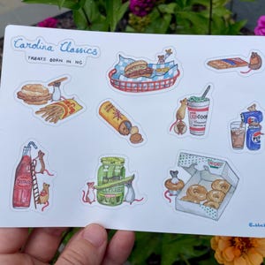 May include: A sticker sheet featuring watercolor illustrations of North Carolina treats. The sheet includes images of a burger, fries, a hotdog, a milkshake, a soda, pickles, donuts, and more. The text "Carolina Classics Treats Born in NC" is at the top.