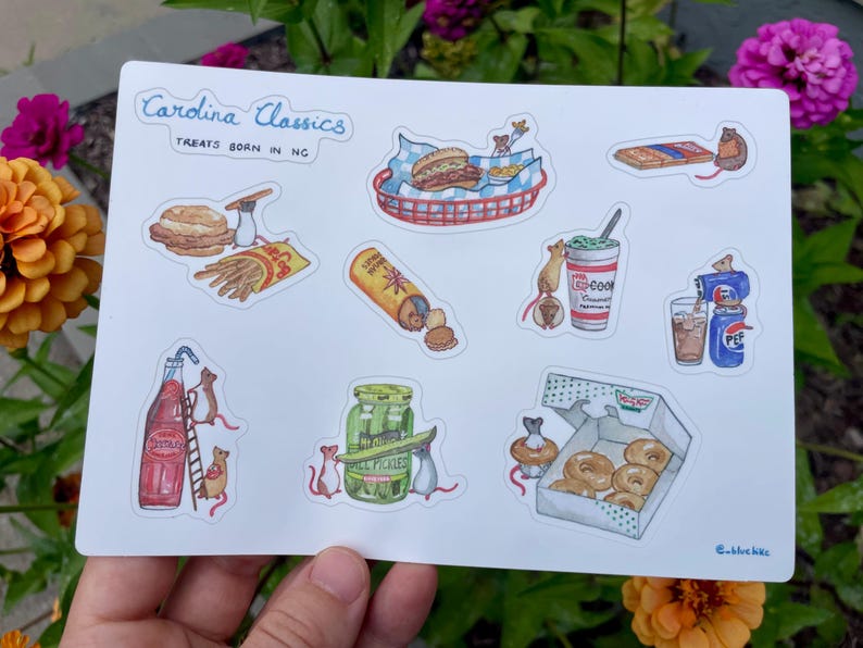 May include: A sticker sheet featuring watercolor illustrations of North Carolina treats. The sheet includes images of a burger, fries, a hotdog, a milkshake, a soda, pickles, donuts, and more. The text "Carolina Classics Treats Born in NC" is at the top.