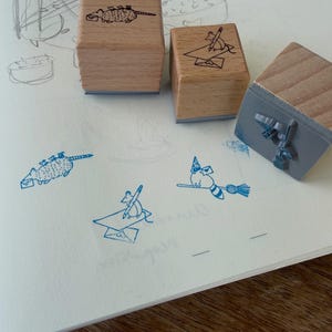 May include: Three square wooden rubber stamps with light brown grain. Two stamps show black ink illustrations of a possum carrying mail and a mouse writing on an envelope. The third stamp is flipped to reveal a gray rubber base with a broom design.