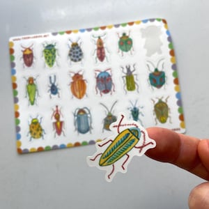 May include: A sheet of colorful insect stickers with a single sticker of a yellow and green beetle with red legs being held by a finger.