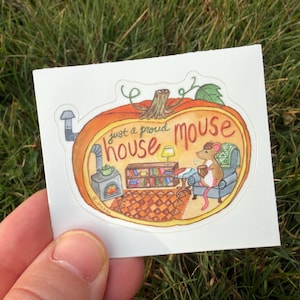May include: A sticker featuring a cozy illustration of a mouse inside a pumpkin. The mouse is seated in a chair, holding a mug, with a fireplace and bookshelf nearby. The text reads "just a proud house mouse."