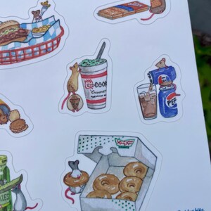 May include: A sheet of watercolor stickers featuring mice interacting with various foods. The stickers include a burger and fries, a milkshake, a soda, a box of donuts, and a package of crackers. The stickers have a whimsical, cartoon style.