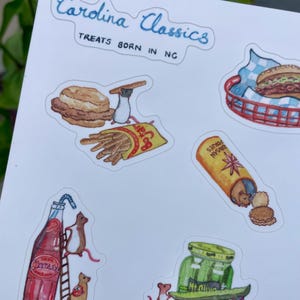 May include: A sticker sheet featuring watercolor illustrations of North Carolina food items. The sheet includes images of a biscuit, french fries, a hot dog, a bottle of Cheerwine, and a jar of pickles. The text "Carolina Classics" and "Treats Born in NC" are also present.