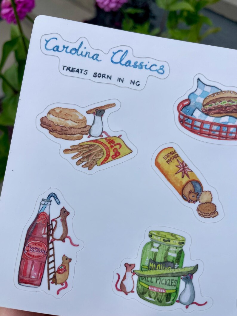May include: A sticker sheet featuring watercolor illustrations of North Carolina food items. The sheet includes images of a biscuit, french fries, a hot dog, a bottle of Cheerwine, and a jar of pickles. The text "Carolina Classics" and "Treats Born in NC" are also present.