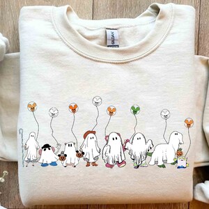 Puede incluir: Una sudadera beige con un gráfico de una línea de fantasmas sosteniendo globos. Los globos son naranja, verde y rojo. Los fantasmas llevan diferentes disfraces, incluyendo un sombrero de bruja, una calabaza y un sombrero de vaquero.