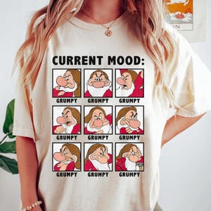 Op de afbeelding: Een wit T-shirt met de tekst "CURRENT MOOD:" en een raster van negen afbeeldingen van Grumpy uit Sneeuwwitje. Elke afbeelding toont een andere gezichtsuitdrukking van Grumpy, en elke afbeelding is gelabeld met "GRUMPY".
