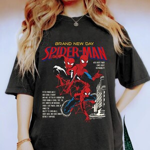 May include: Black t-shirt featuring a Spider-Man graphic. The design includes the words "BRAND NEW DAY" and "SPIDER-MAN" in red and white, along with illustrations of Spider-Man in action. Text details Peter Parker's origin and powers.