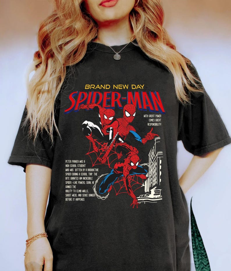 May include: Black t-shirt featuring a Spider-Man graphic. The design includes the words "BRAND NEW DAY" and "SPIDER-MAN" in red and white, along with illustrations of Spider-Man in action. Text details Peter Parker's origin and powers.