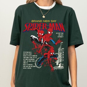 May include: Dark green t-shirt featuring a Spider-Man graphic. The design includes the words "BRAND NEW DAY" and "SPIDER-MAN" in red and blue, along with comic book illustrations of Spider-Man. The shirt also has text describing Spider-Man's origin.