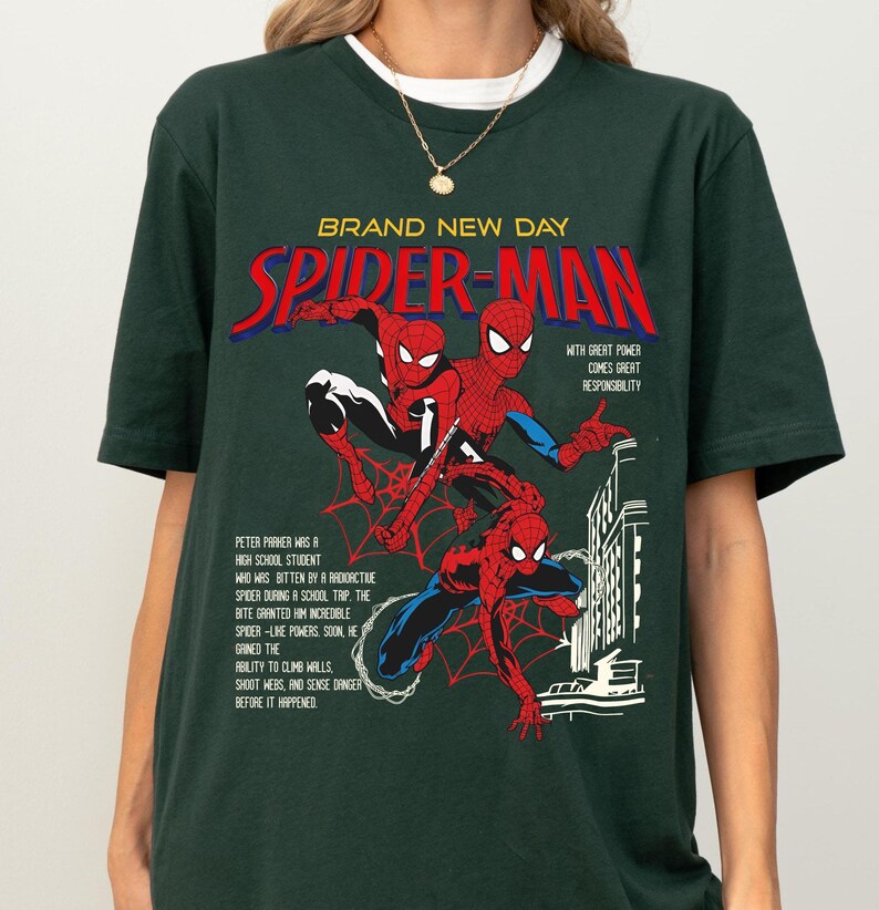 May include: Dark green t-shirt featuring a Spider-Man graphic. The design includes the words "BRAND NEW DAY" and "SPIDER-MAN" in red and blue, along with comic book illustrations of Spider-Man. The shirt also has text describing Spider-Man's origin.