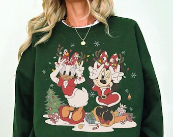 May include: A dark green crewneck sweatshirt featuring a graphic print of Daisy Duck and Minnie Mouse wearing red holiday outfits and reindeer antlers. The design includes a small Christmas tree, wrapped gifts, and white snowflakes.