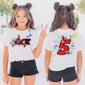 Pu&ograve; includere: T-shirt bianca con il nome "Jack" in stile fumetto, con grafica di Spider-Man. Il retro della maglietta mostra un grande numero rosso "5" con un disegno a ragnatela e personaggi di Spider-Man. La maglietta &egrave; indossata da una persona giovane.