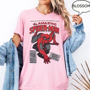 May include: Pink t-shirt featuring a comic book graphic of Spider-Man in red and black. The text "the AMAZING SPIDER-MAN" is displayed above the character. Additional text includes "NEVER HAS ANYONE FOUGHT SUCH MERCILESS FOES AS..." and "BLOSSOM".