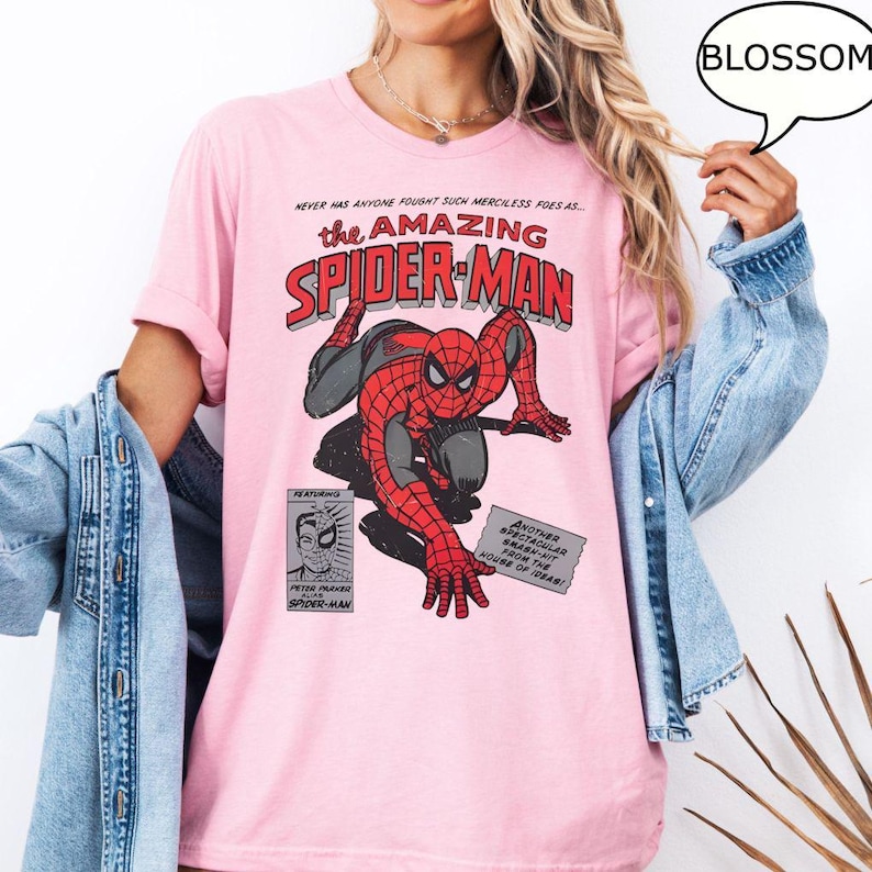 May include: Pink t-shirt featuring a comic book graphic of Spider-Man in red and black. The text "the AMAZING SPIDER-MAN" is displayed above the character. Additional text includes "NEVER HAS ANYONE FOUGHT SUCH MERCILESS FOES AS..." and "BLOSSOM".