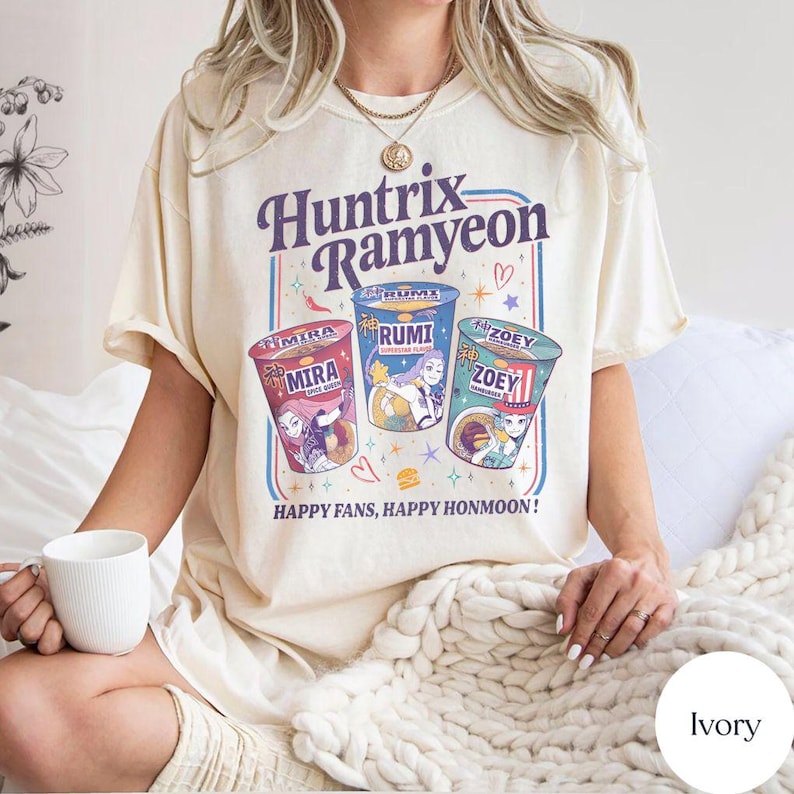 Puede incluir: Camiseta de color marfil con el texto "Huntrix Ramyeon" sobre tres dise&ntilde;os ilustrados de tazas de ramen. Cada taza es de un color diferente y presenta un personaje &uacute;nico. El texto "Happy Fans, Happy Honmoon!" est&aacute; debajo.