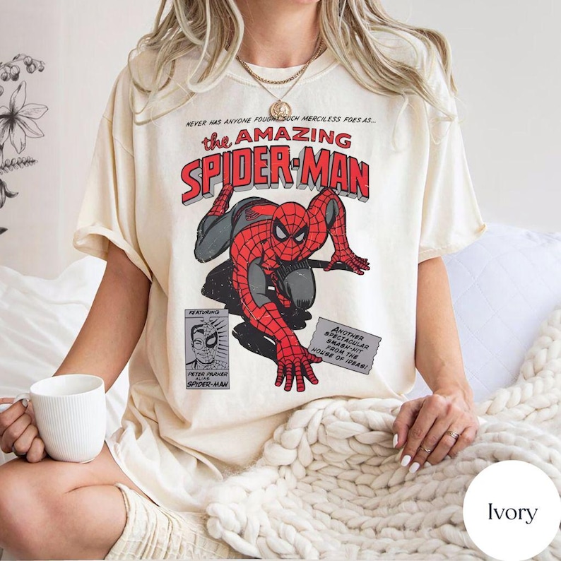 May include: Ivory-colored t-shirt featuring a vintage comic book design of Spider-Man. The graphic includes the words "the AMAZING SPIDER-MAN" in red and black, with Spider-Man in a classic pose. The shirt also has additional text and a small image of Peter Parker.