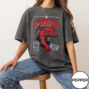 May include: Dark gray t-shirt featuring a comic book graphic of Spider-Man in red and black. The text "The Amazing Spider-Man" is displayed in red and white. The shirt has a vintage, worn look.