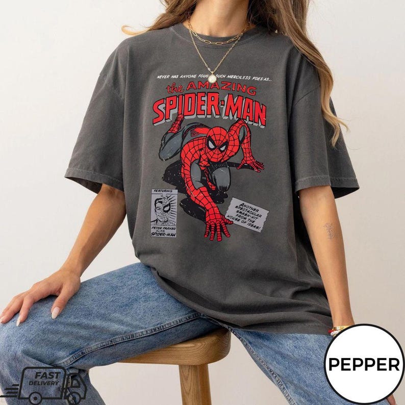 May include: Dark gray t-shirt featuring a comic book graphic of Spider-Man in red and black. The text "The Amazing Spider-Man" is displayed in red and white. The shirt has a vintage, worn look.