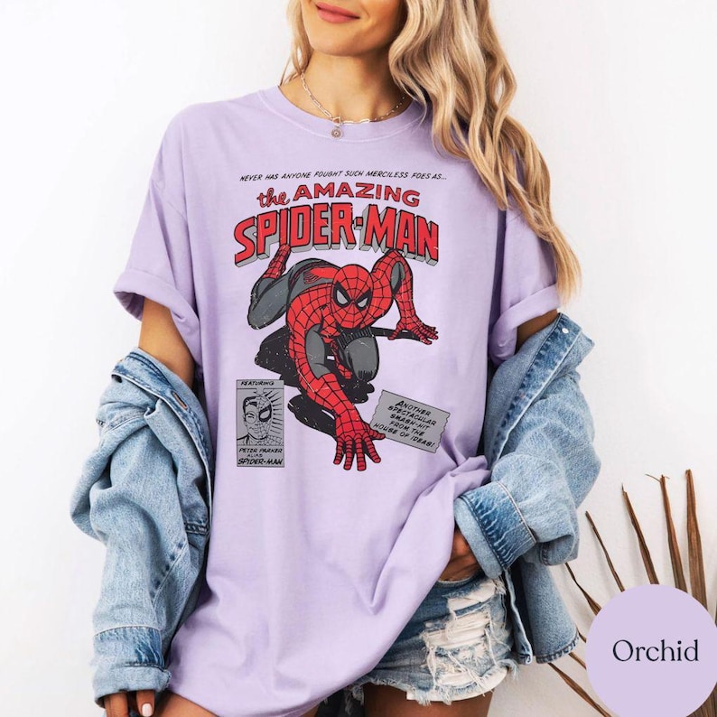 May include: Orchid-colored t-shirt featuring a comic book graphic of Spider-Man in a dynamic pose. The design includes the text "the AMAZING SPIDER-MAN" in red and white, along with comic book panels. The shirt is a casual, short-sleeved style.