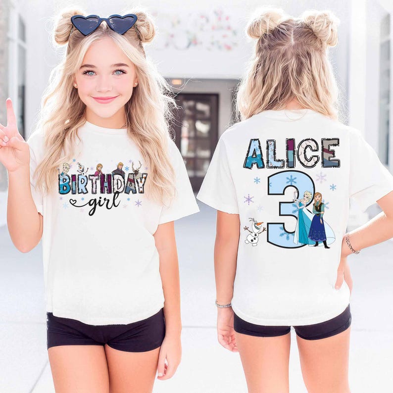 May include: A white short-sleeve t-shirt featuring a graphic print. The front displays the text "BIRTHDAY girl" with cartoon characters and snowflakes. The back shows the name "ALICE" above a large number "3" with cartoon characters and snowflakes.