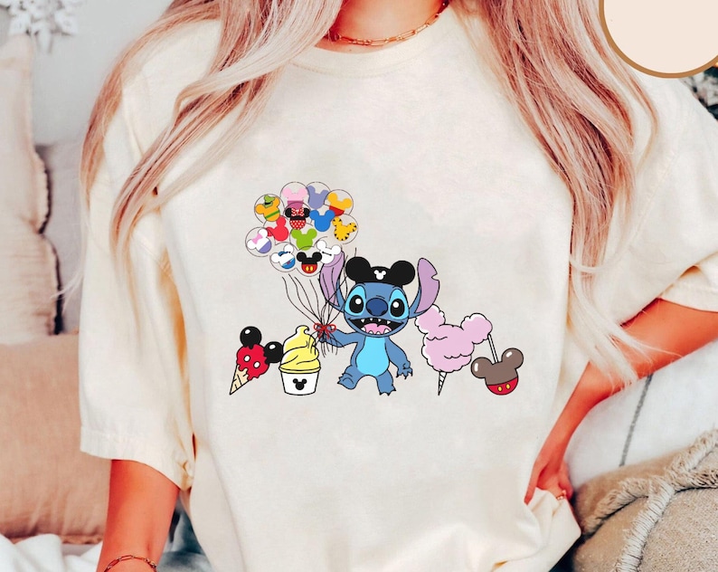 May include: A white t-shirt with a blue Stitch character holding balloons with different Disney characters on them. Stitch is wearing a black Mickey Mouse hat. There are also Mickey Mouse ears, a Mickey Mouse ice cream cone, and a Mickey Mouse cotton candy.
