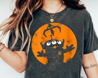 May include: A charcoal grey short-sleeve t-shirt featuring a graphic of a three-eyed alien character beneath a claw machine crane, set against a distressed orange circular background. The alien has its arms raised and wears a suit with a planet emblem.