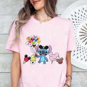 May include: Pink t-shirt featuring a blue Stitch character holding a bunch of balloons with Mickey Mouse faces. The balloons are surrounded by other Disney characters, including Mickey Mouse, Minnie Mouse, Donald Duck, and Goofy. The shirt also features a Mickey Mouse ice cream cone, a cotton candy, and a Mickey Mouse ear hat.