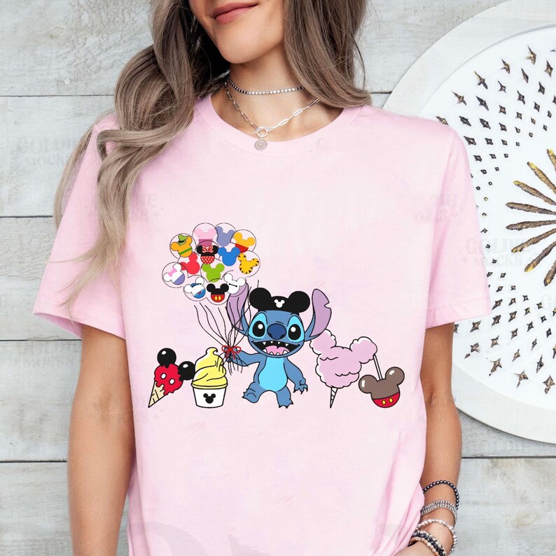 May include: Pink t-shirt featuring a blue Stitch character holding a bunch of balloons with Mickey Mouse faces. The balloons are surrounded by other Disney characters, including Mickey Mouse, Minnie Mouse, Donald Duck, and Goofy. The shirt also features a Mickey Mouse ice cream cone, a cotton candy, and a Mickey Mouse ear hat.